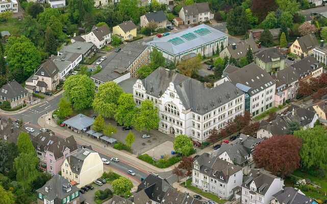 Zu sehen ist ein Luftbild des Nikolaus-Ehlen-Gymnasiums. Mittig in dem Foto liegt das Gebäude des Gymnasiums, im Vordergrund der Schulhof, die Bushaltestelle sowie die Friedrich-Ebert-Straße. Im Hintergrund ist die Sporthalle der Schule sowie das umliegende Wohngebiet zu sehen.