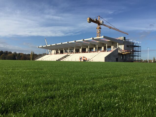 im Hintergrund eine Stadiontribüne im Bau zu sehen, ein Baukran, im Vordergrund Rasen