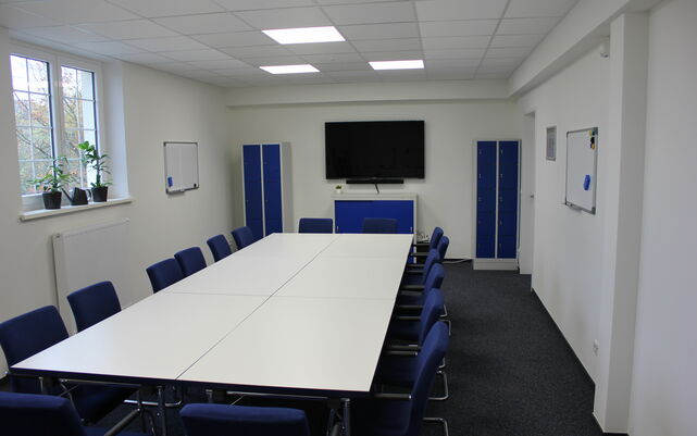 Innenaufnahme mit langem Konferenztisch und blauen Stühlen im Vordergrund, Fernseher/ Beamer im Hintergrund.