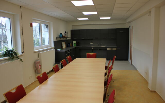 Innenaufnahme mit langem Konferenztisch und roten Stühlen im Vordergrund, im Hintergrund die graue Küche.