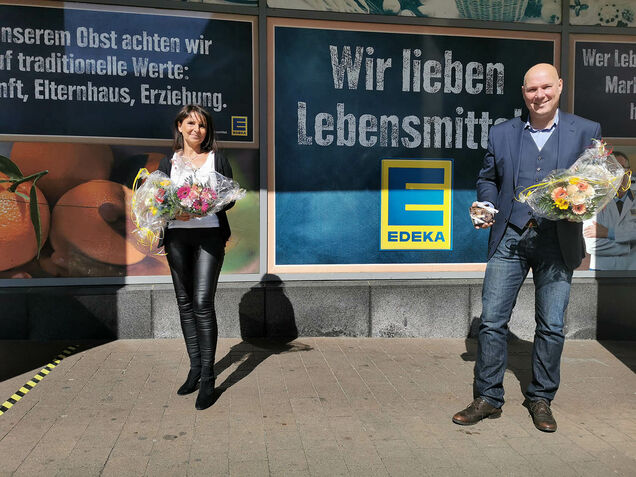 Zu sehen sind Bürgermeister Dirk Lukrafka und eine städtische Mitarbeiterin mit je einem Blumenstrauß in der Hand vor dem Eingang von Edeka.